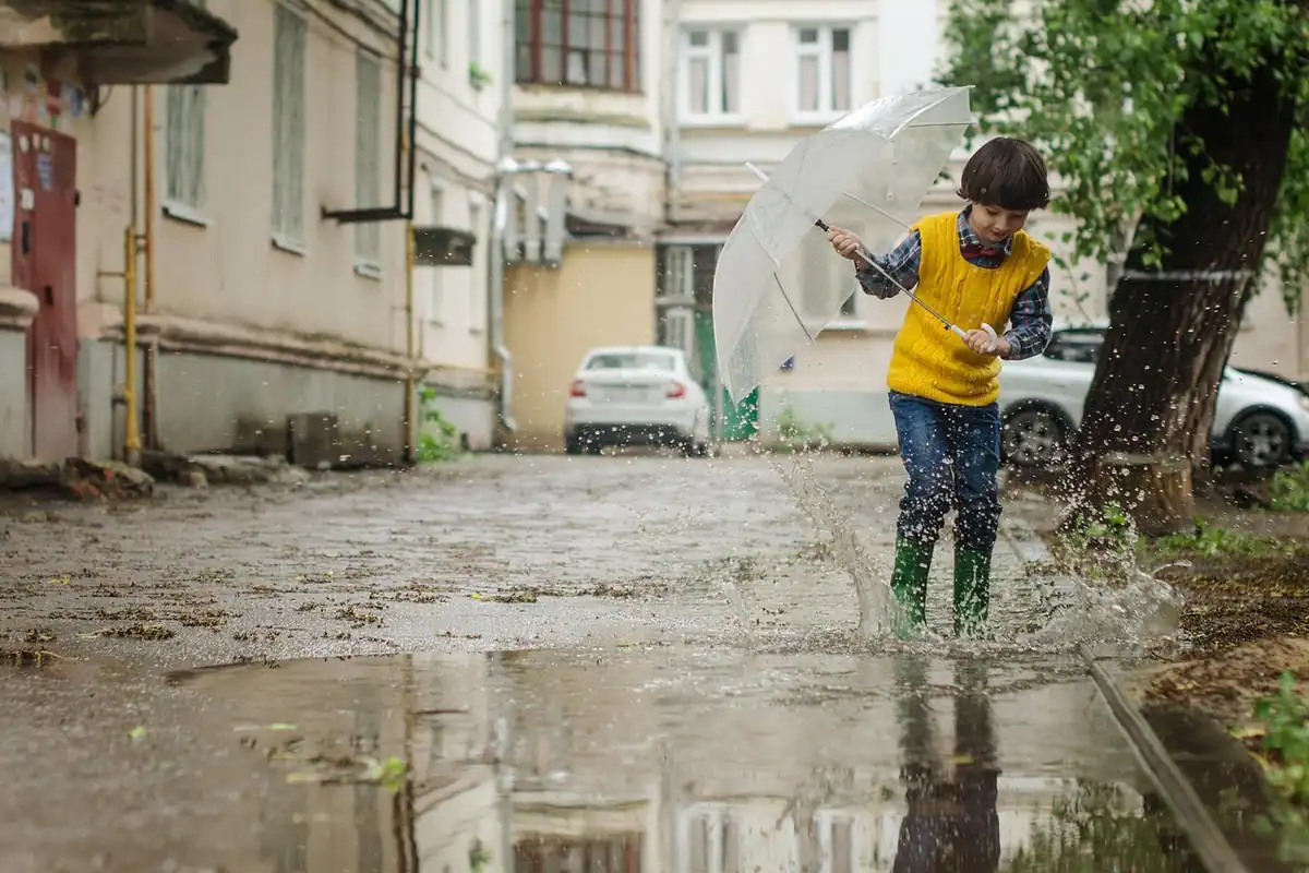Băiat în cizme de cauciuc care sare printr-o băltoacă, pe o stradă inundată între blocuri