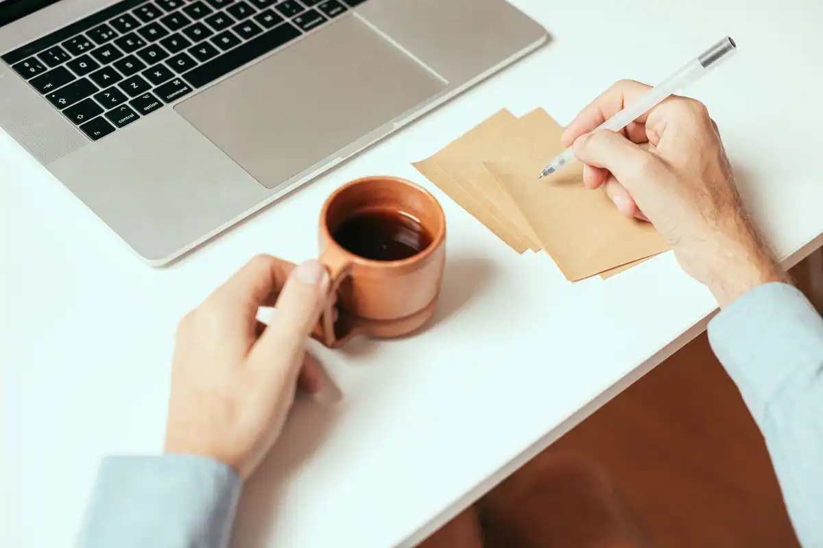 Mâini care scriu cu un creion pe bilețele, lângă un laptop și o ceașcă de cafea