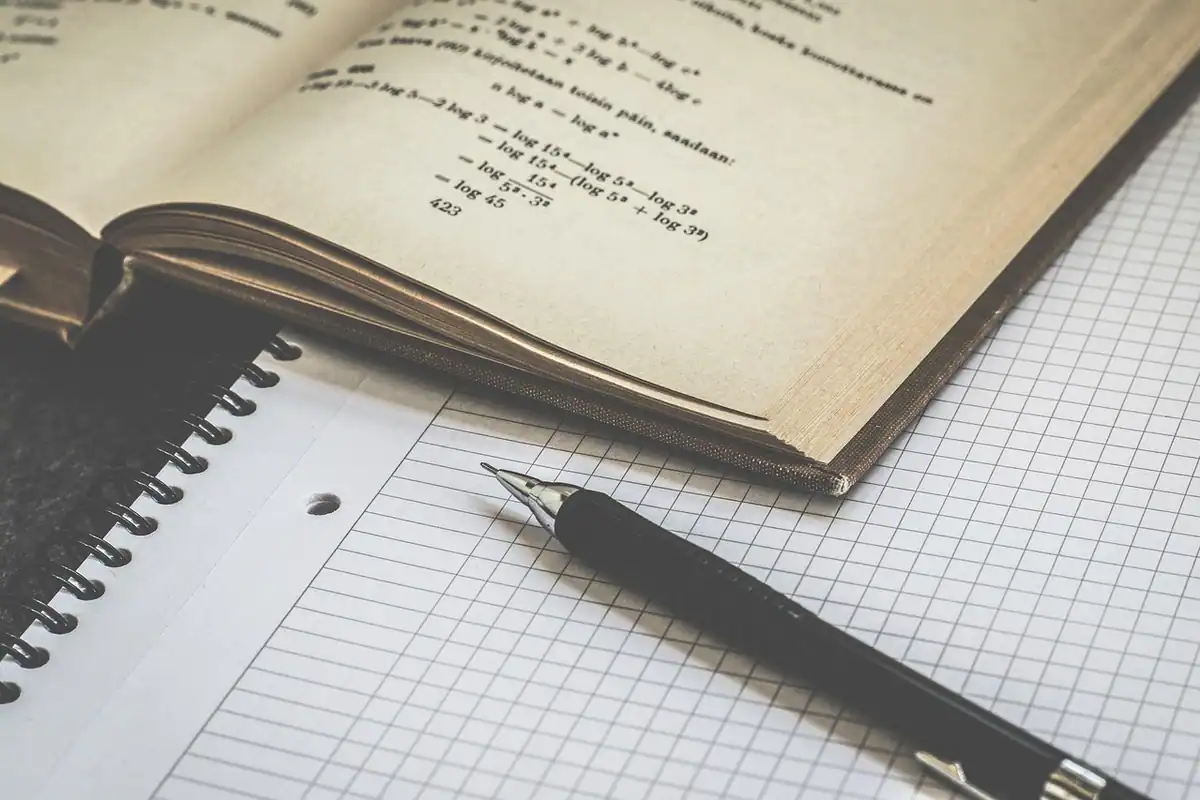 Caiet cu pătrățele și stilou lângă o carte deschisă cu formule, pentru calculul punctajului de pensie