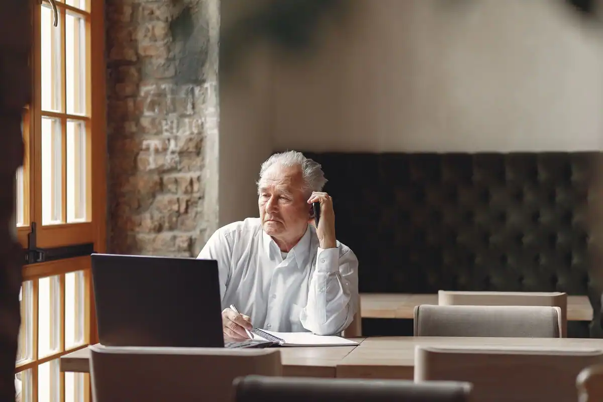Bărbat în vârstă care lucrează la laptop, la o masă de lângă ferestre luminoase