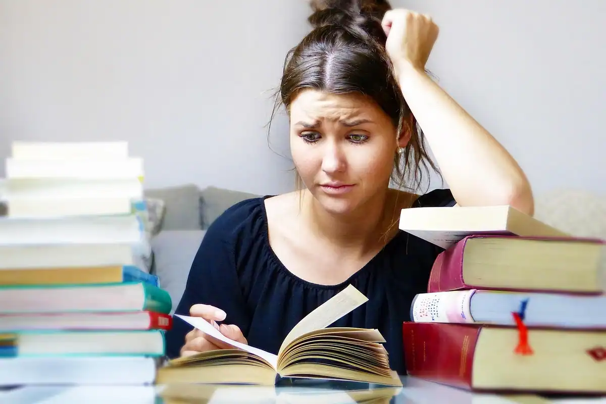 Tânără obosită între teancuri de cărți, care învață pentru examene și se gândește la costurile studiilor