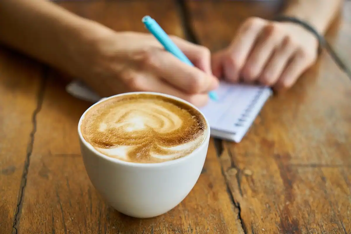 Mână care notează cu un pix într-un carnet, lângă o ceașcă de cafea cu spumă