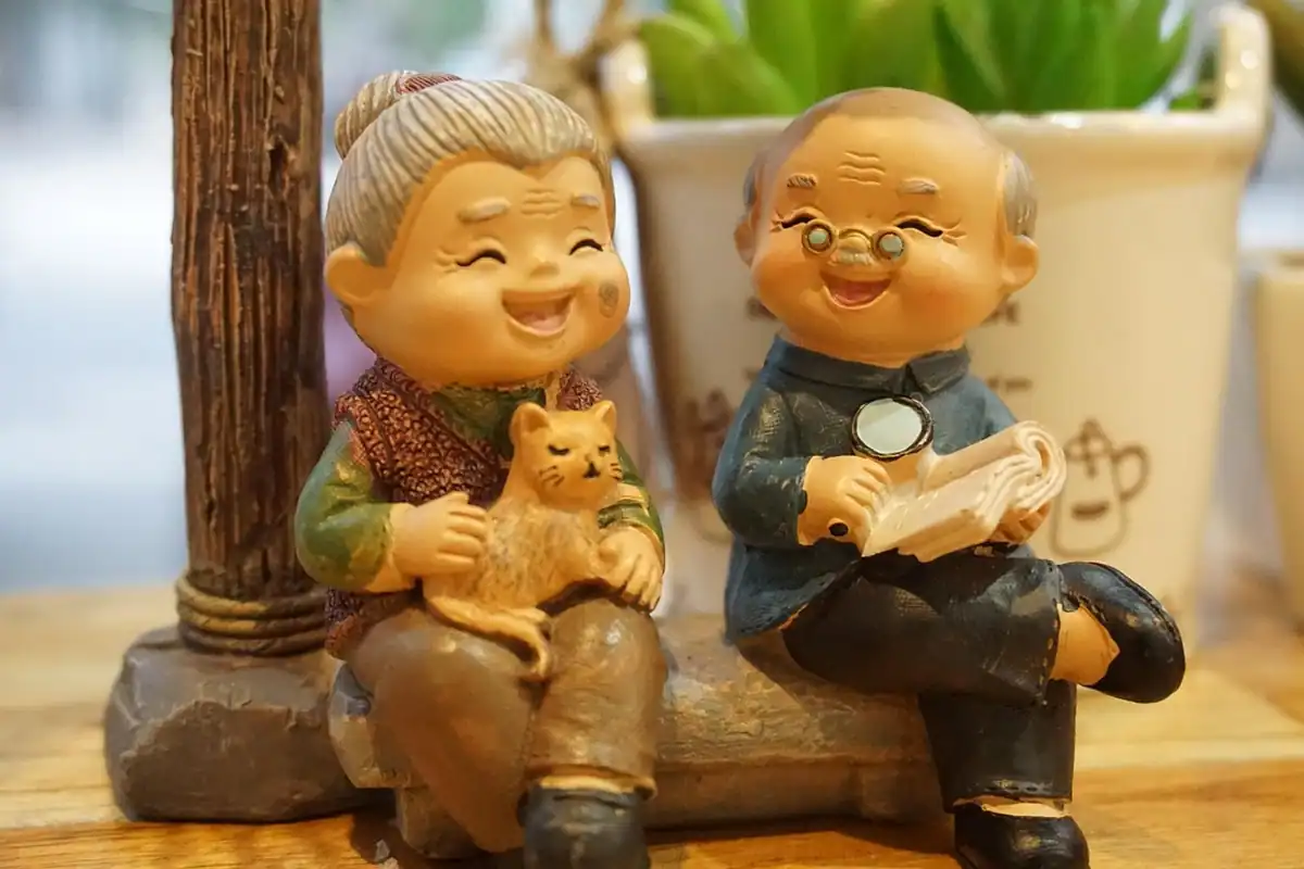 Figurine care înfățișează două persoane în vârstă zâmbitoare, cu o pisică și o carte
