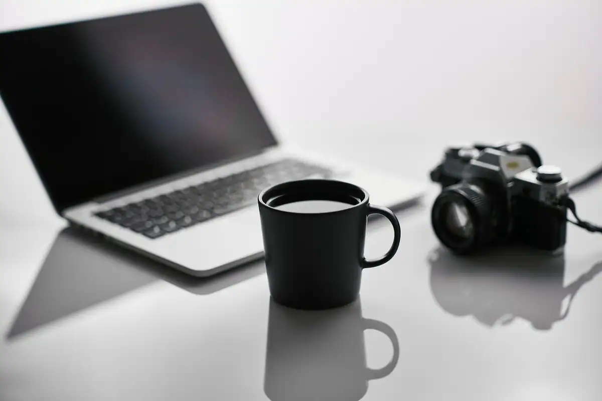 Laptop, cană neagră și aparat foto pe un birou alb, locul de muncă al unui liber-profesionist
